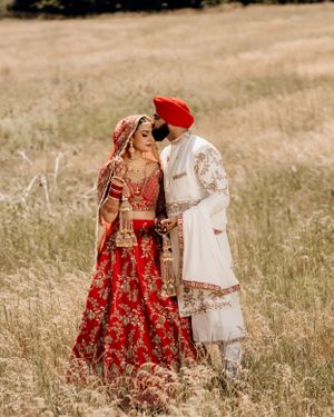 Sikh wedding couple in traditional red and white attire pose in a golden field.