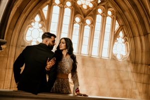 Elegant wedding photo with couple against gothic arched windows in warm lighting.
