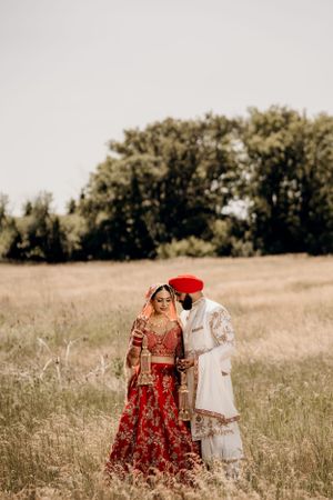 Artistic wedding portrait of couple in traditional Indian attire standing in a rustic field.
