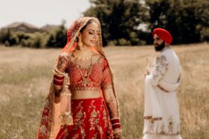 Wedding portrait showing bride in red lehenga with groom in white sherwani in background.