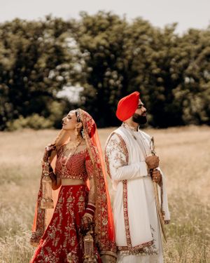 A couple in traditional Indian wedding attire poses together in a rustic outdoor field setting.