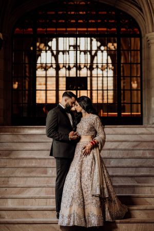 Couple shares intimate moment on grand stairs in front of ornate gothic window during wedding photoshoot.