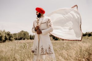 White sherwani outfit with red turban photographed in sunlit field with flowing fabric.