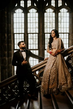 A romantic couple poses on an ornate gothic staircase with arched windows in formal attire.