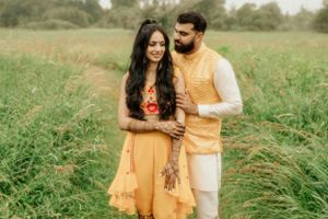 Couple in matching yellow and white traditional Indian outfits pose together in a green field.