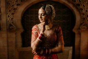 Bride in red lehenga poses against ornate archway in moody lighting.
