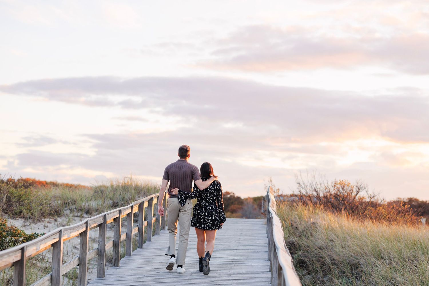 A couple walks along a wooden boardwalk path through beach dunes during a scenic sunset.