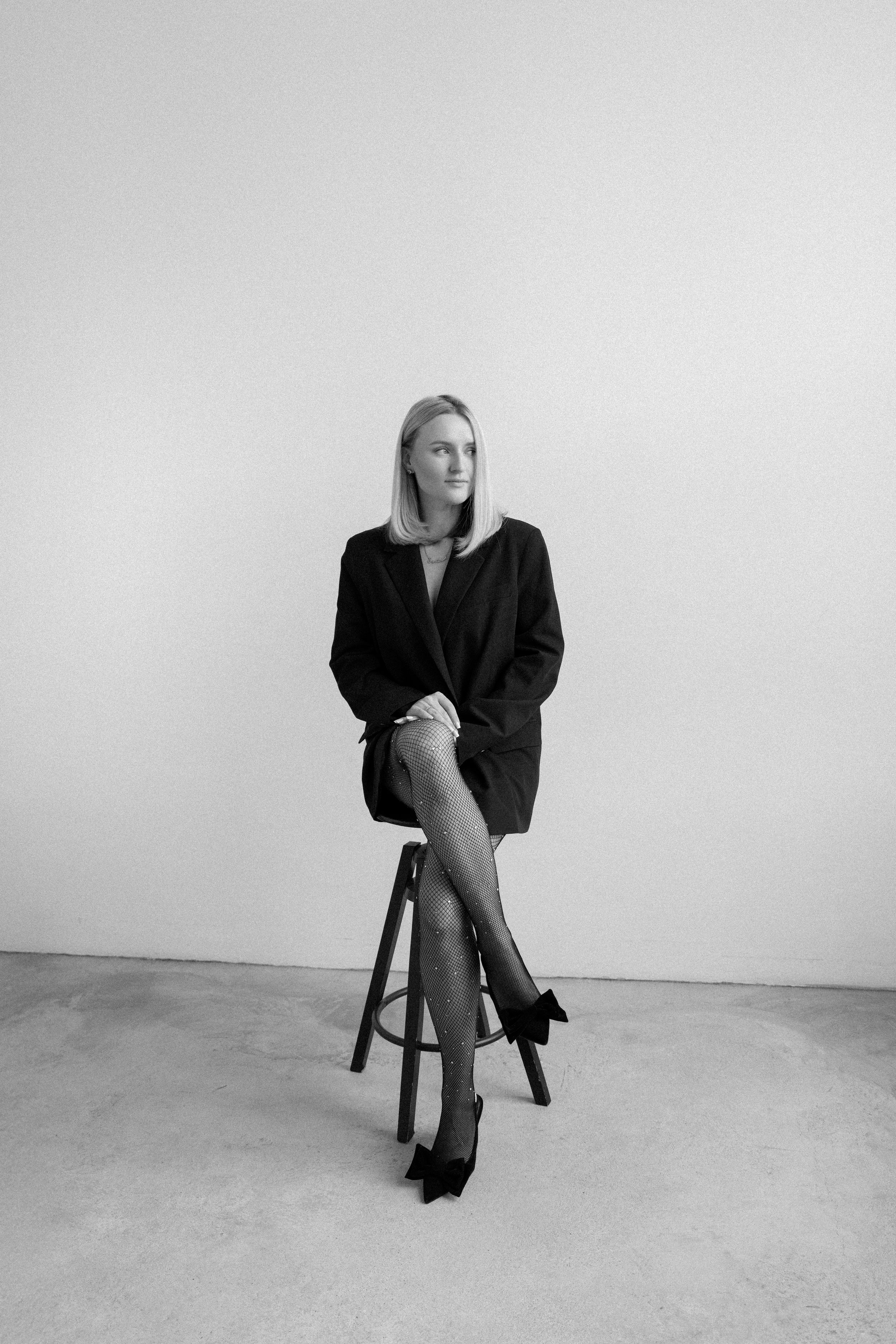 Black and white portrait of woman in blazer seated on stool with crossed legs and high heels.