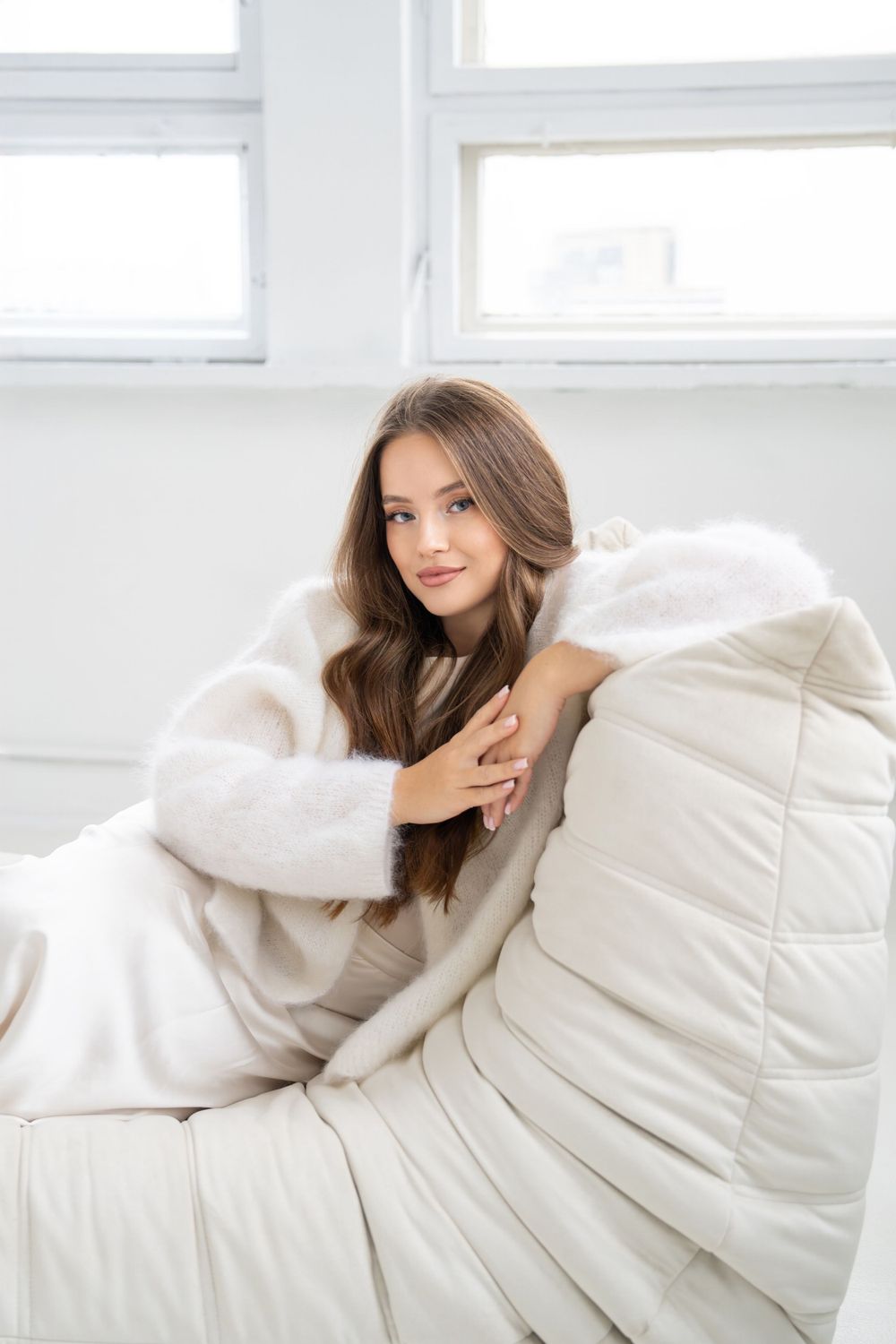 Woman in cozy white sweater poses on plush white couch near window.