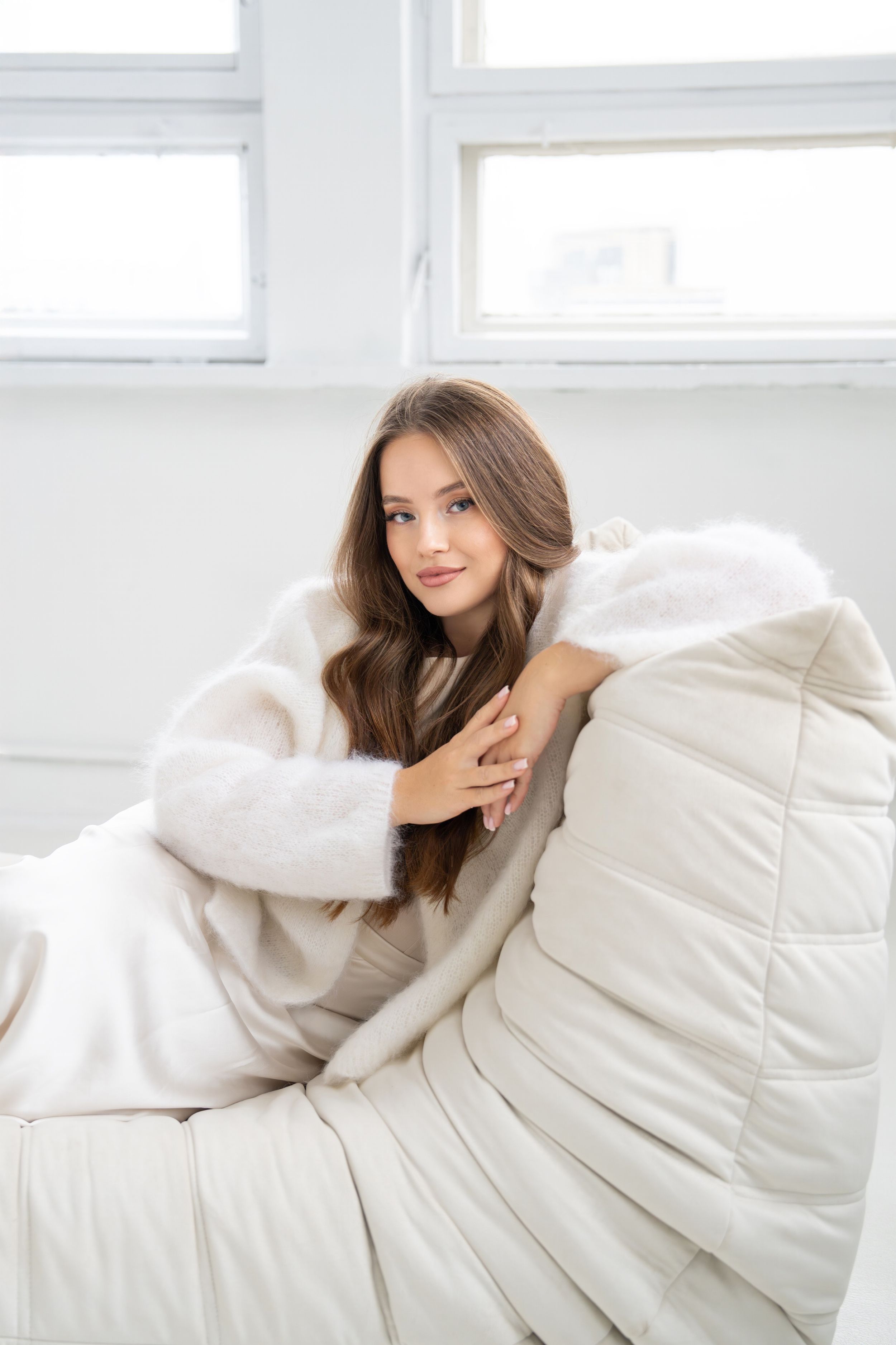 Woman in cozy white sweater poses on plush white couch near window.