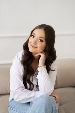 A person in a white button-down shirt and jeans poses casually on a light gray couch in a minimalist setting.