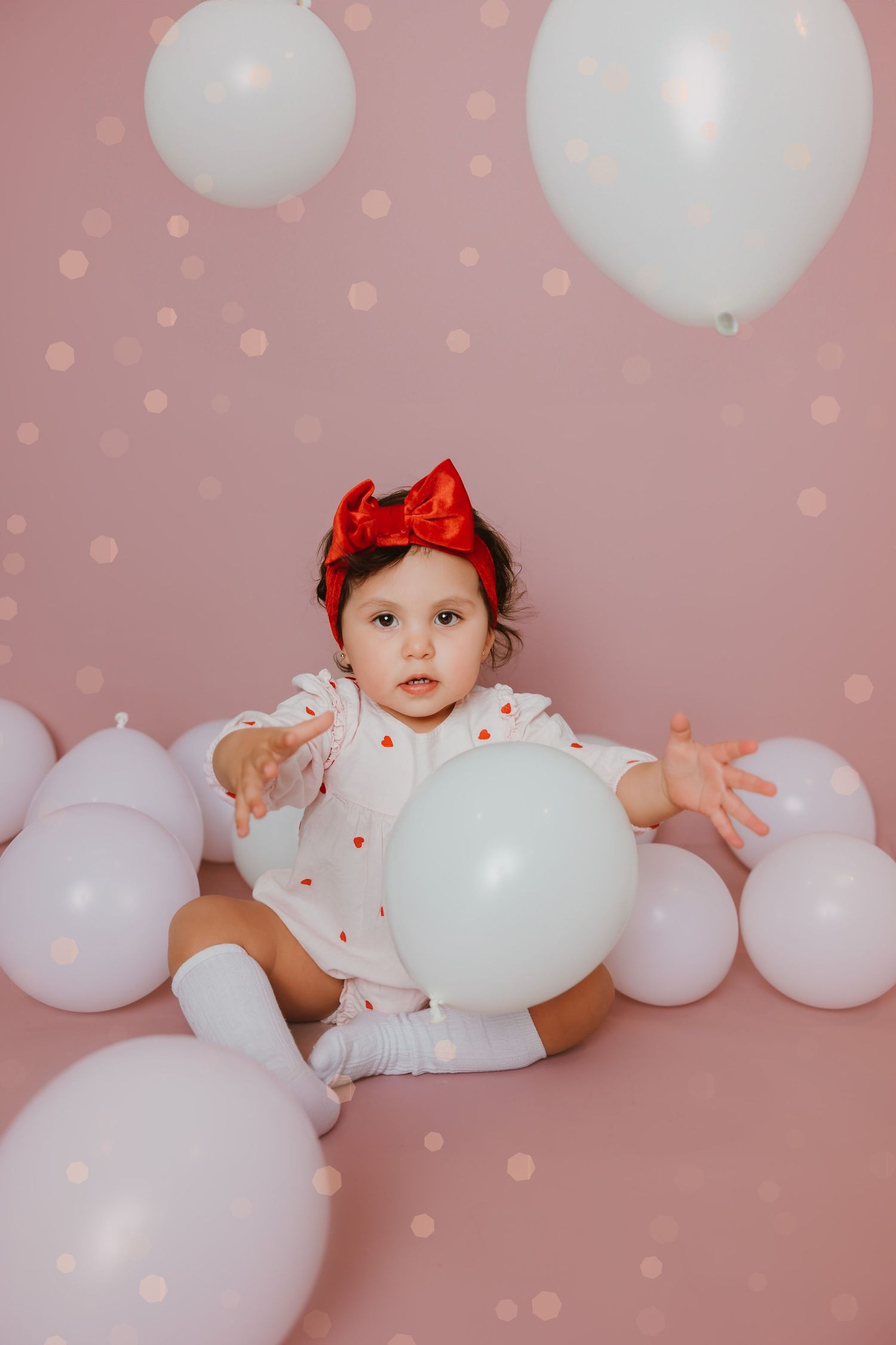 Baby in white dress with red bow surrounded by white balloons on pink backdrop.