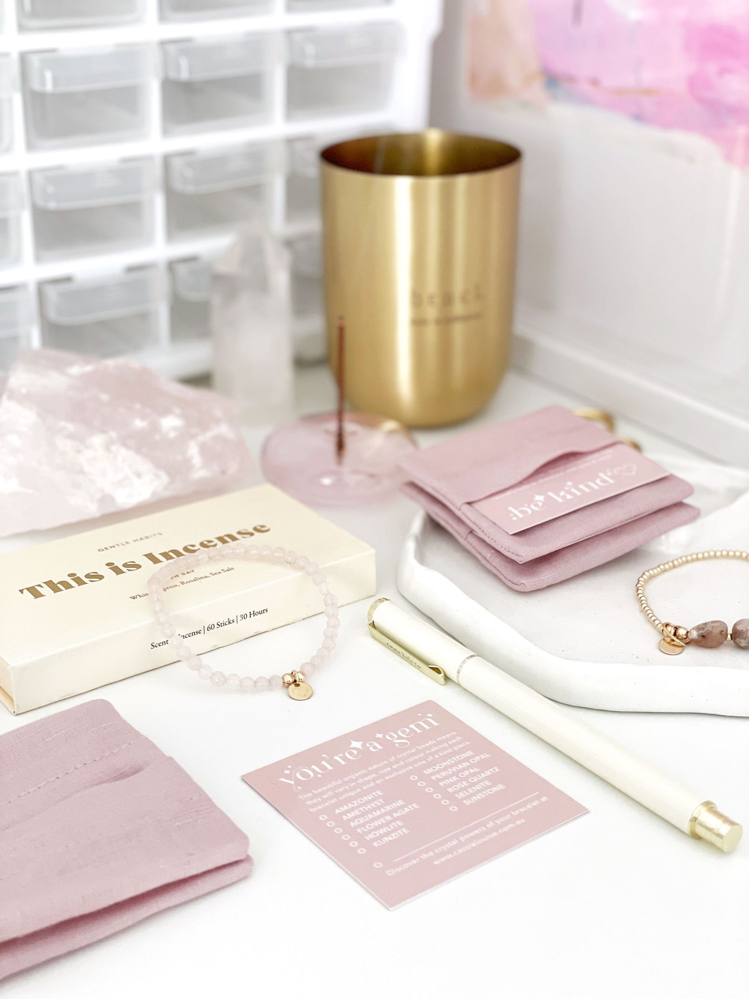 Elegant desktop setup with gold tumbler, pink stationery items and delicate jewelry on white surface.