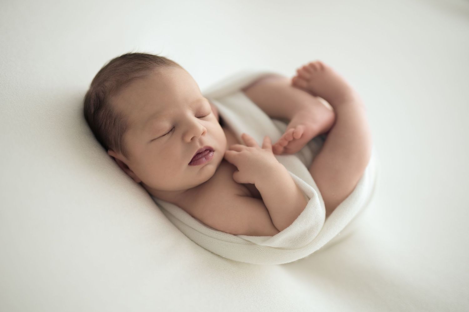 Neonato dorme rilassato su un pouf bianco avvolto in una fasciatura morbida e in una posa naturale