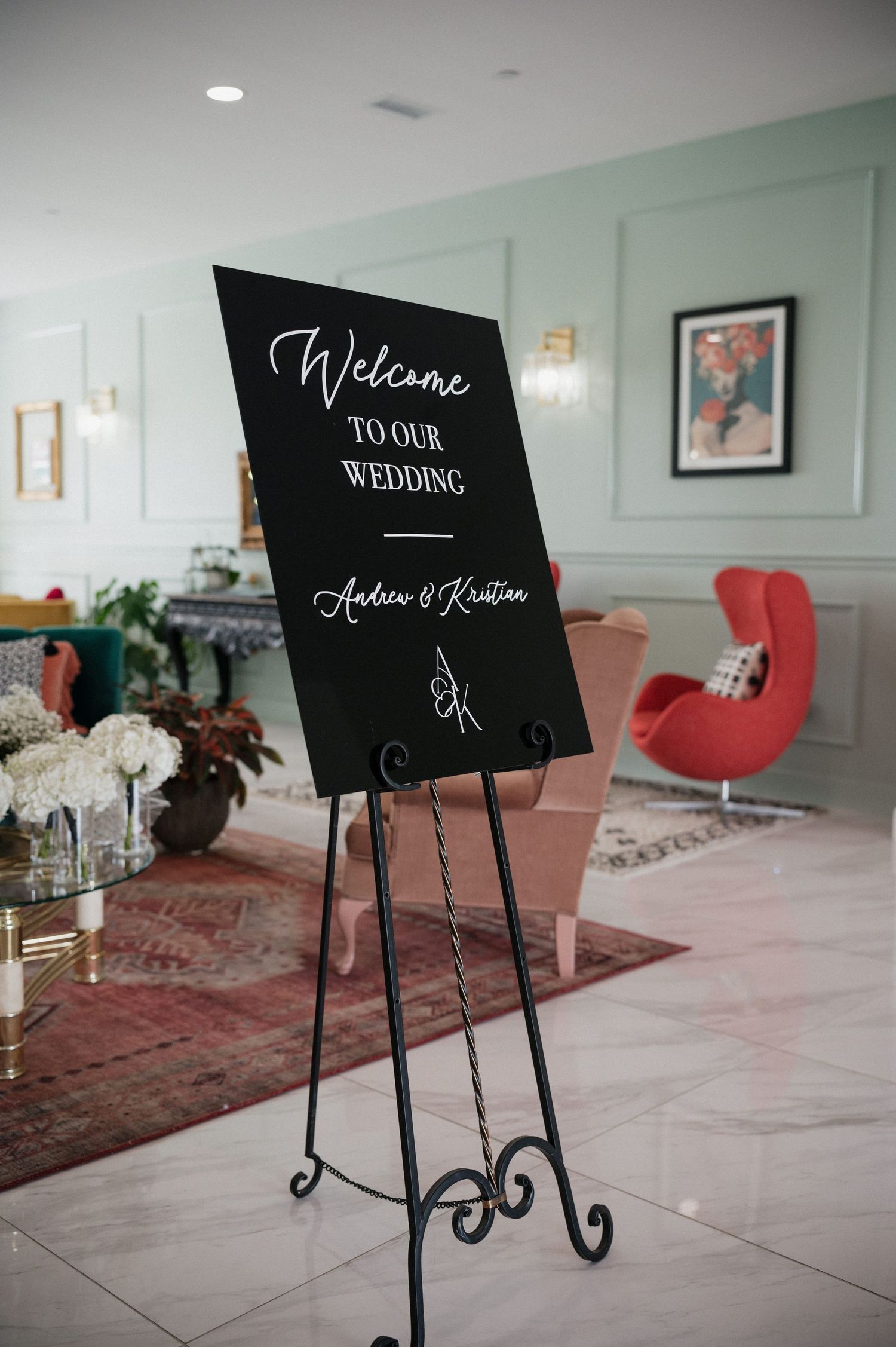 Black wedding welcome sign on an ornate metal easel stand with a modern red chair and vintage rug in the background.