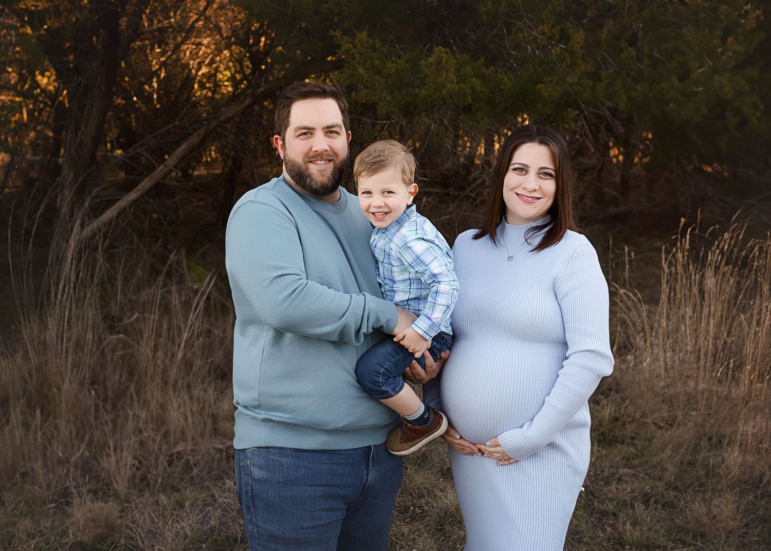 Outdoor maternity photo of family in casual sweaters against autumn foliage background.