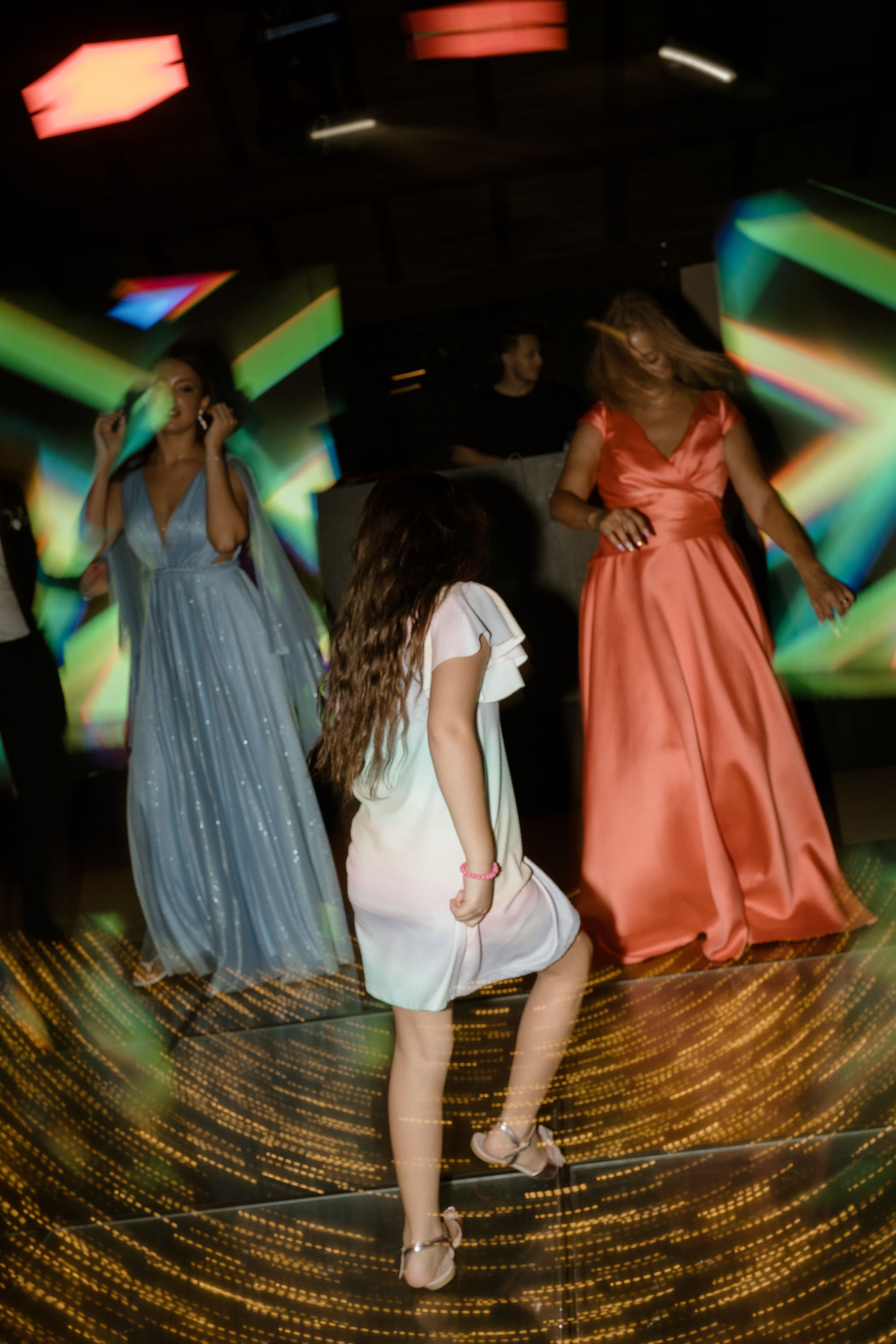 Women in elegant dresses dance under colorful disco lights at a party.