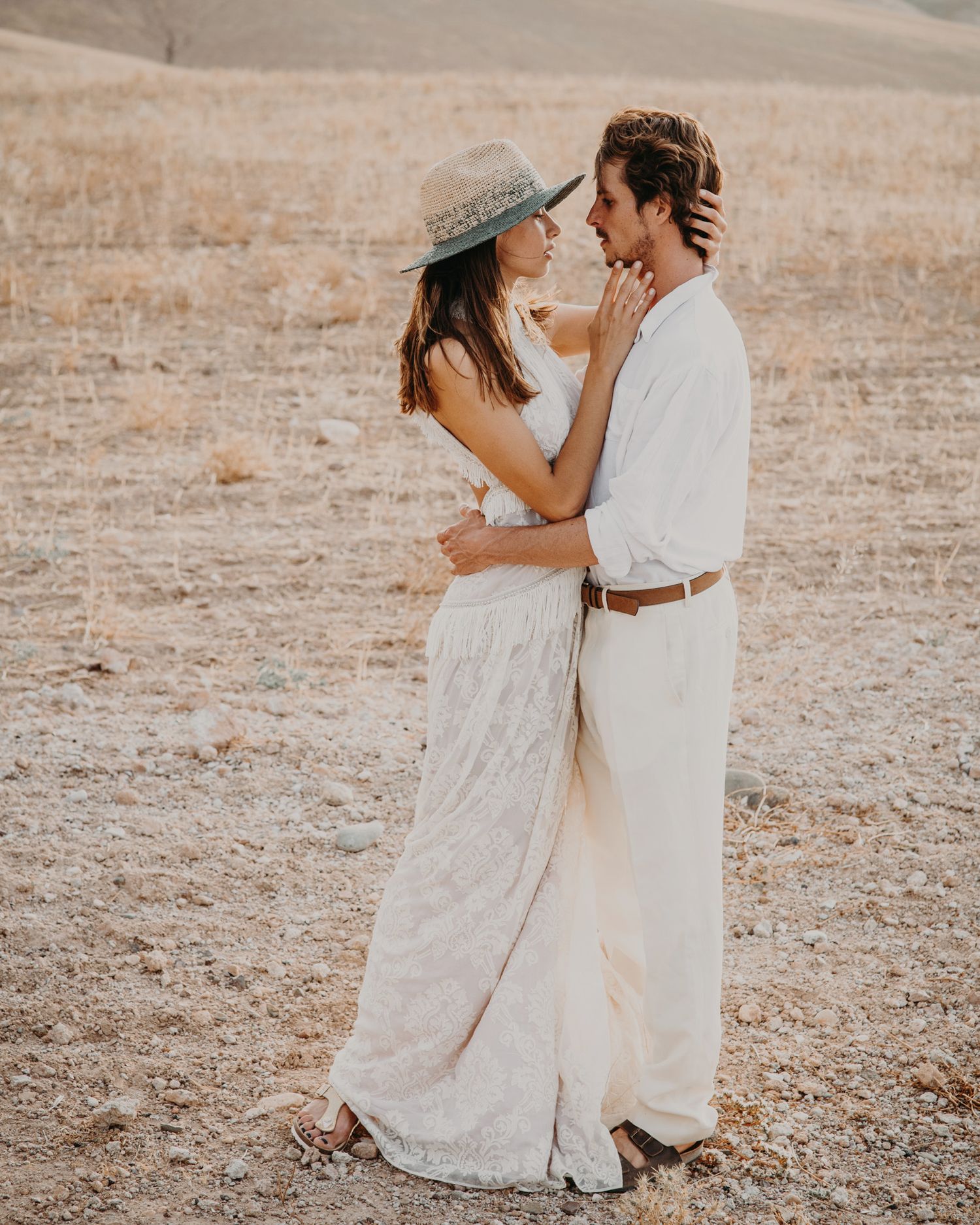 Romantic couple in white outfits embracing on a sun-dried field with a vintage hat.