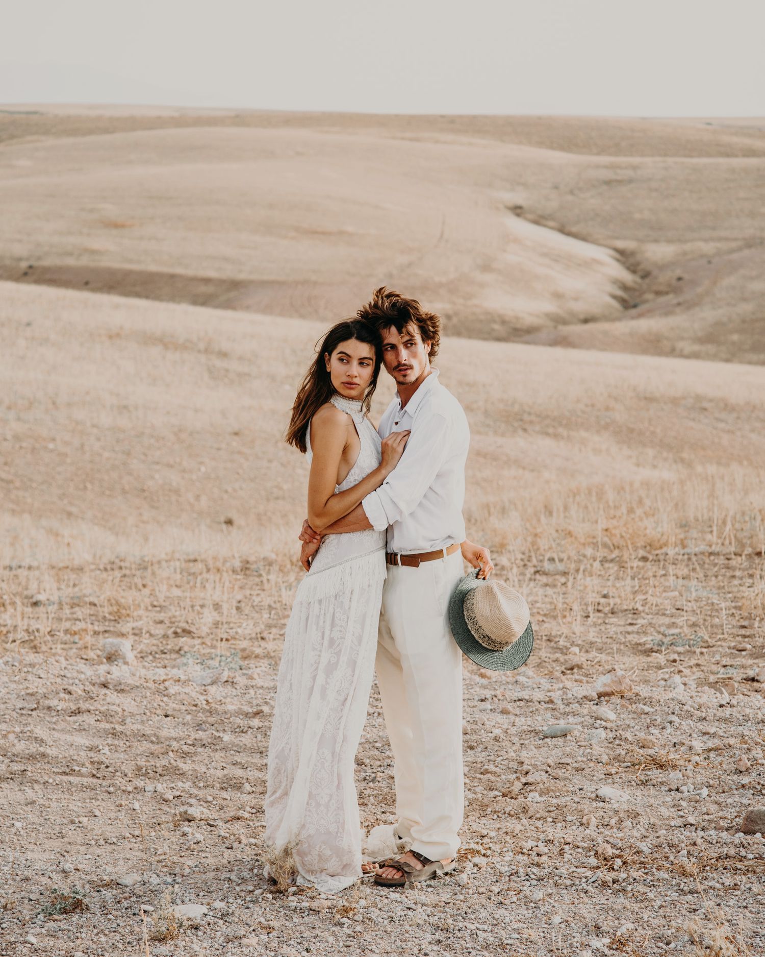 Couple in ethereal white clothing embrace in a sun-bleached desert landscape.
