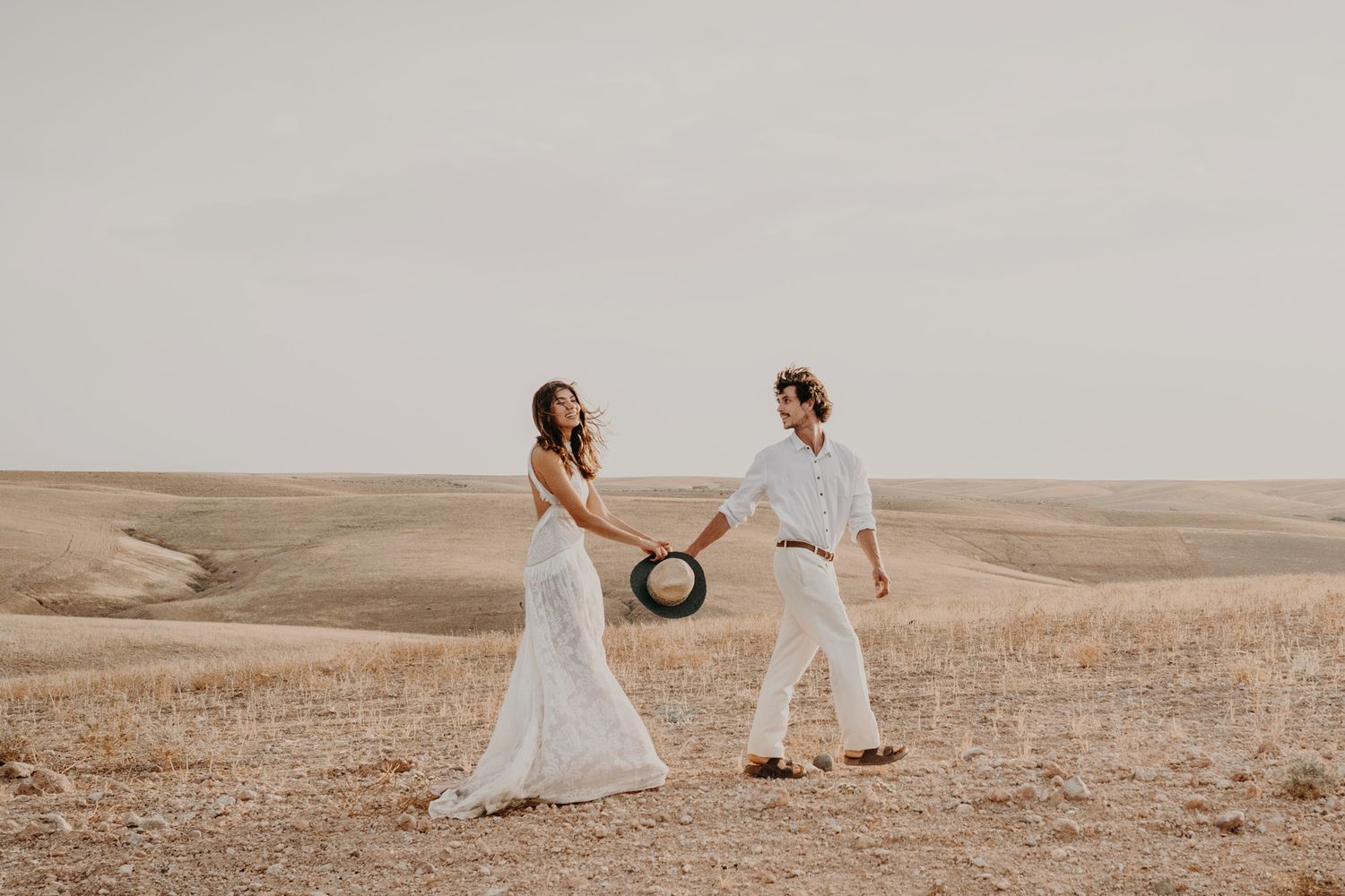 Dreamy desert photoshoot of a couple in white attire holding a vintage hat.