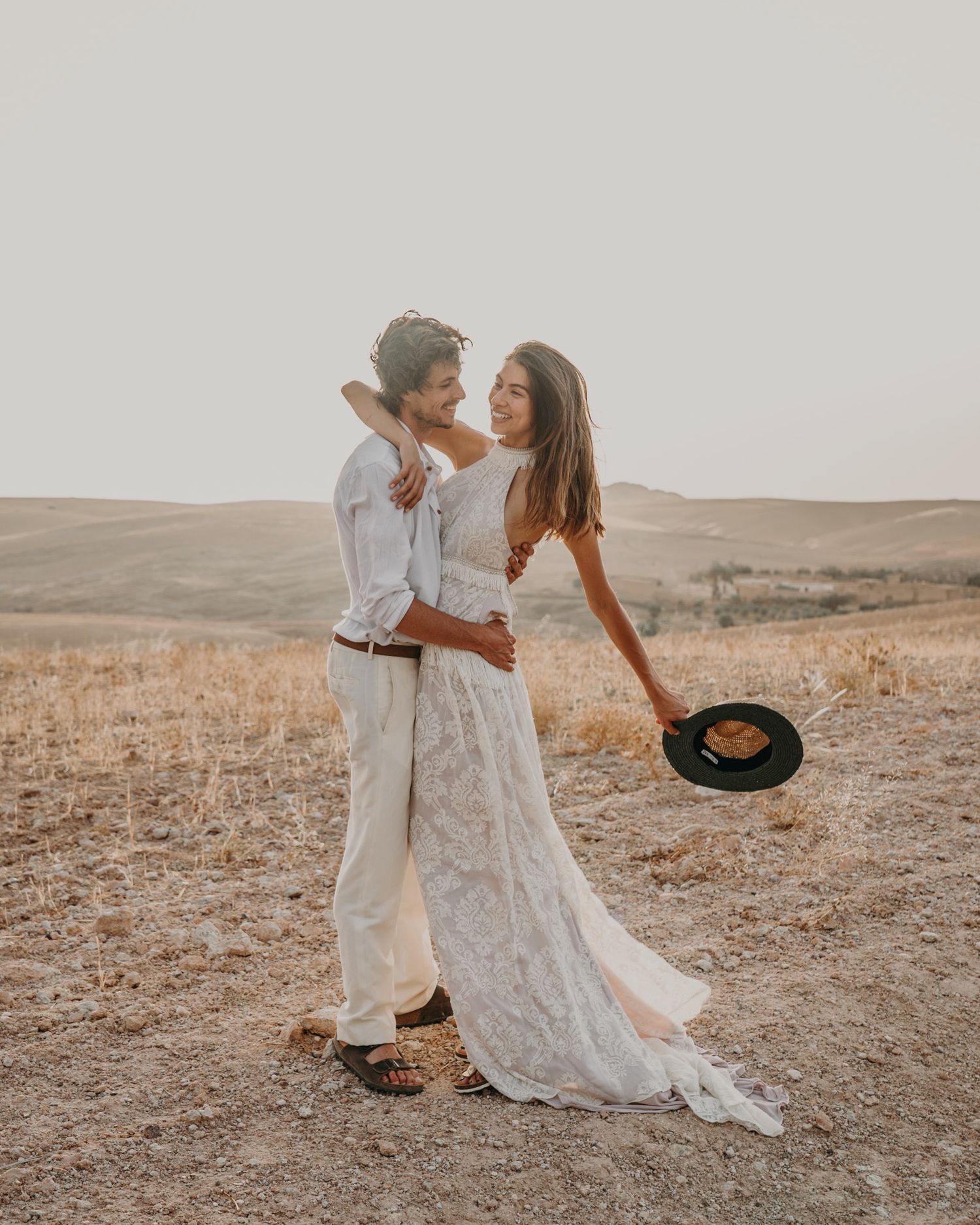 Romantic couple in flowing white outfits share a playful moment in a desert setting.