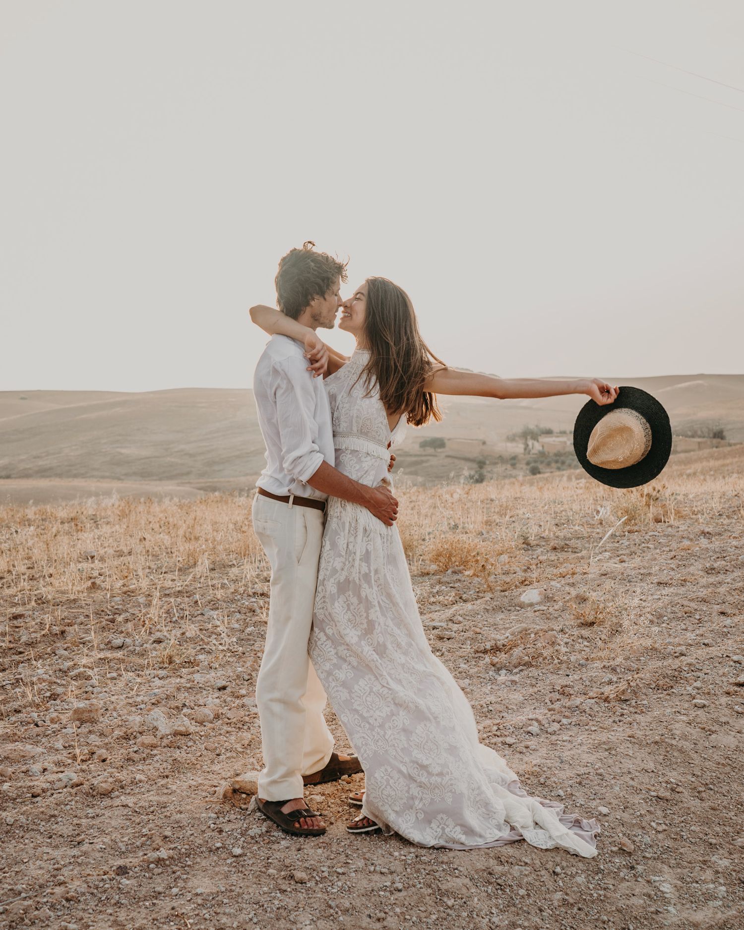Couple in white clothes shares a romantic embrace while holding a vintage hat in a desert.