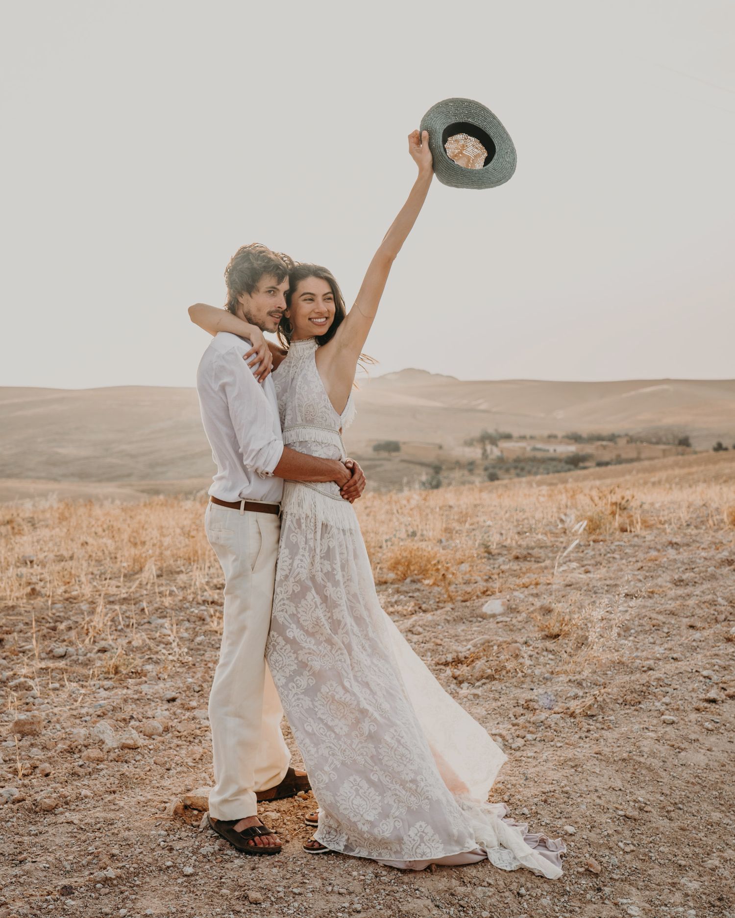 Joyful couple in white celebrates with raised hat against desert backdrop.