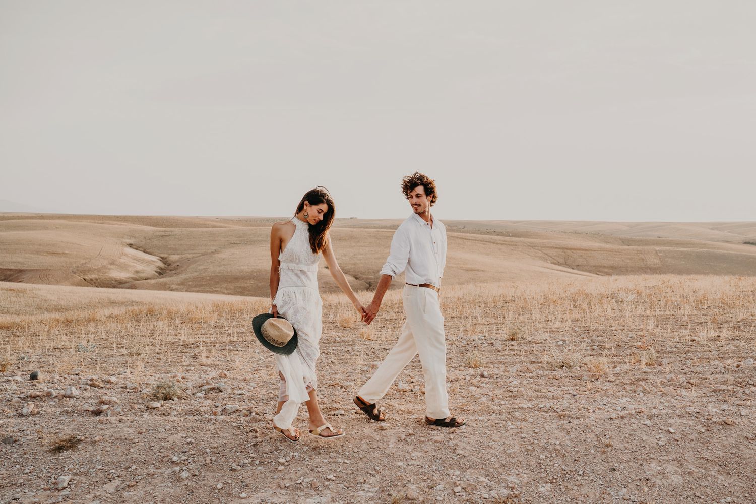 A romantic couple walking hand in hand through a golden field at sunset wearing light colored outfits.
