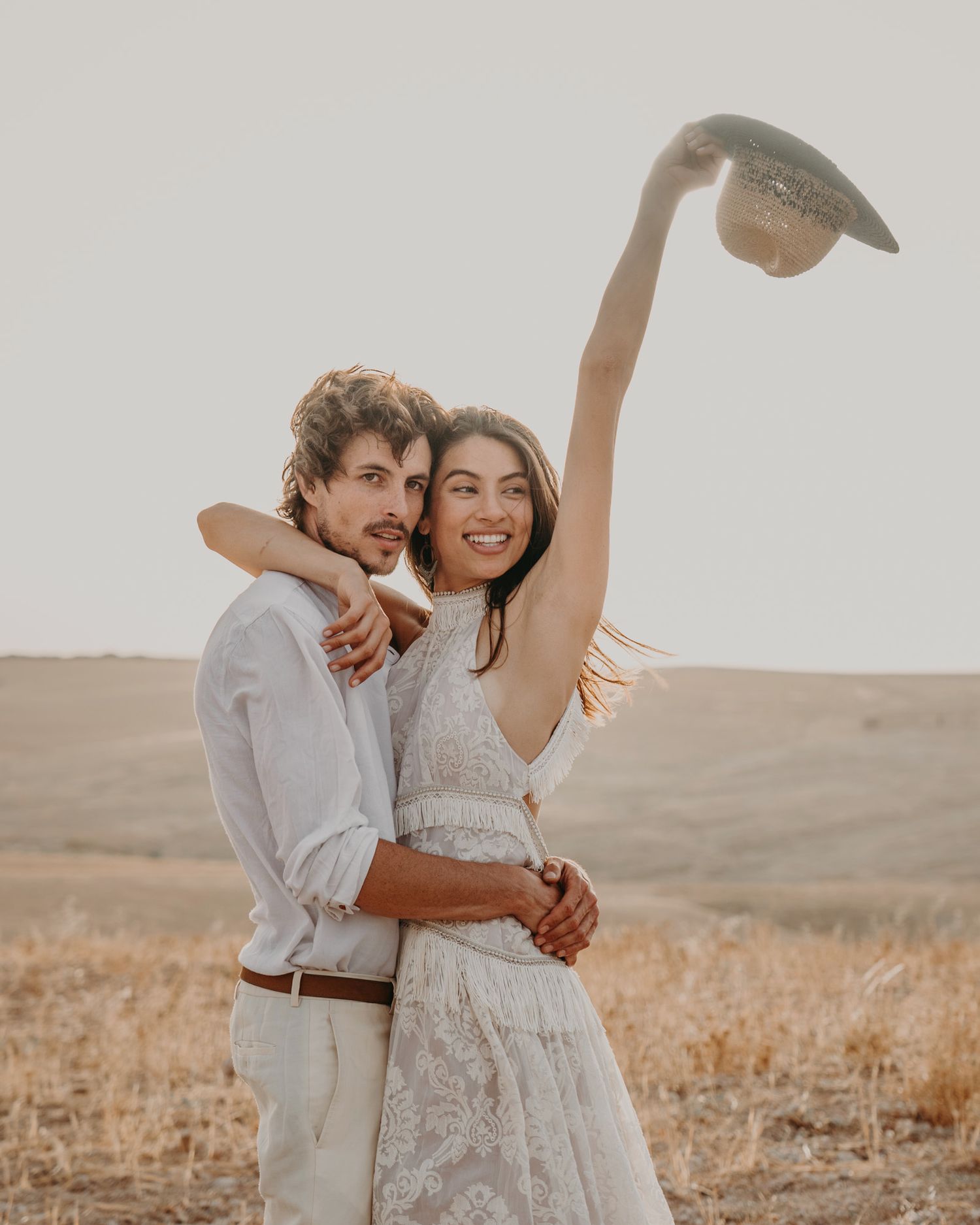 A joyful couple embracing in a field at golden hour while holding up a hat.