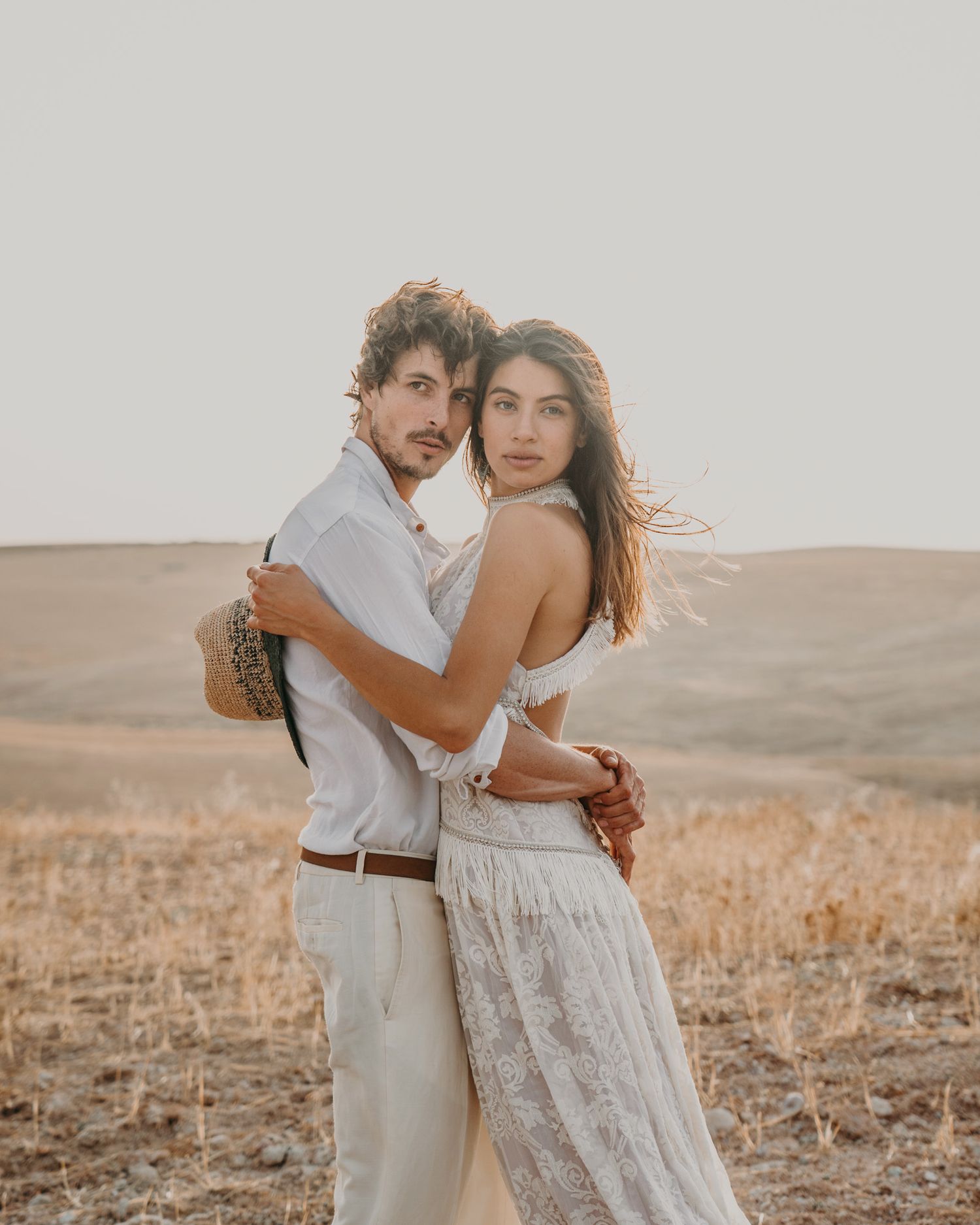 A couple in light colored clothing embracing in a desert landscape at sunset.