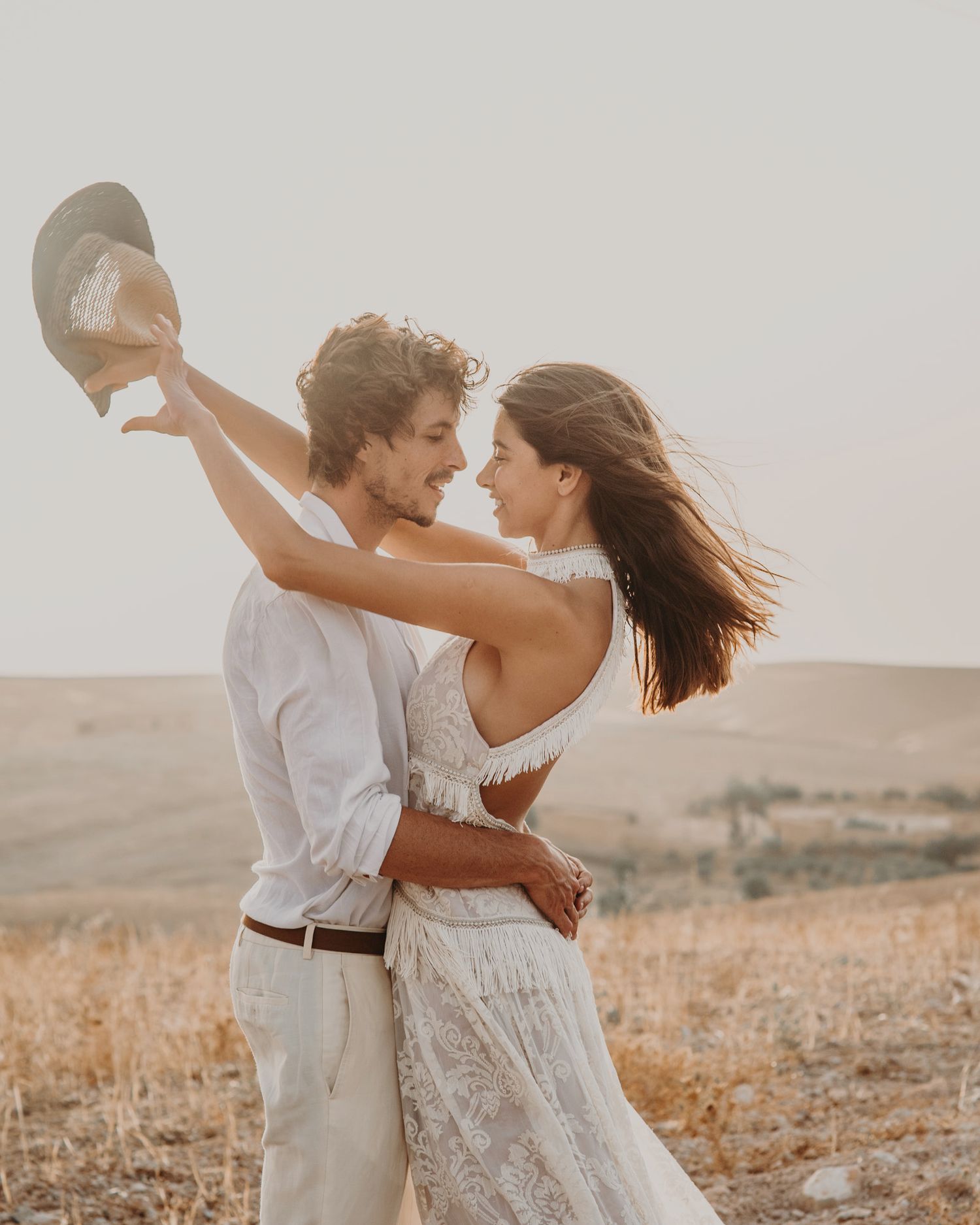 A couple dancing and twirling with a hat in a golden field at sunset.
