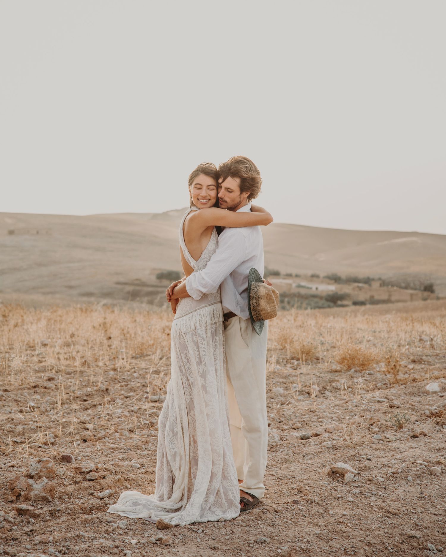 A loving embrace between a couple in white clothing against rolling desert hills.