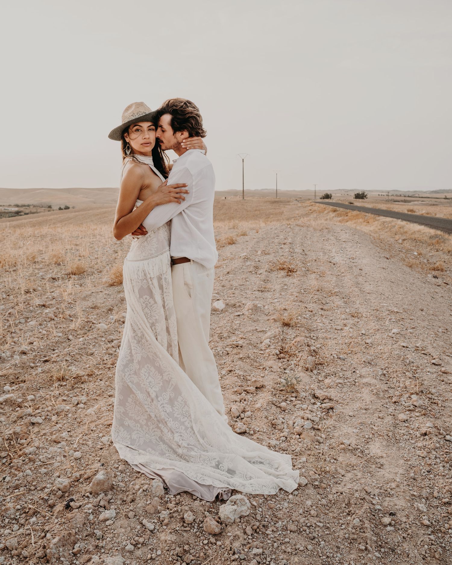 A couple embraces on a desert road wearing flowing white wedding attire.