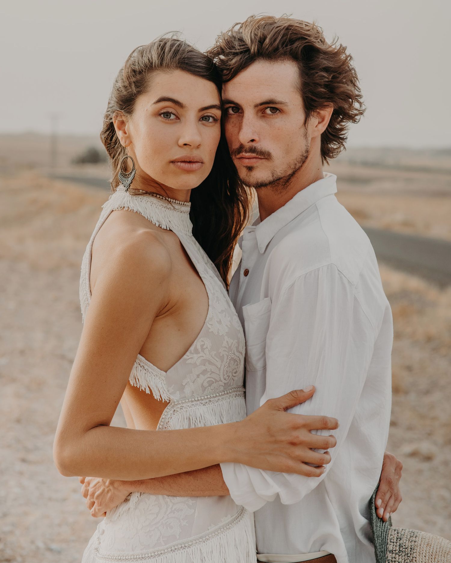 A romantic desert engagement photo of a couple in white clothing standing close together.