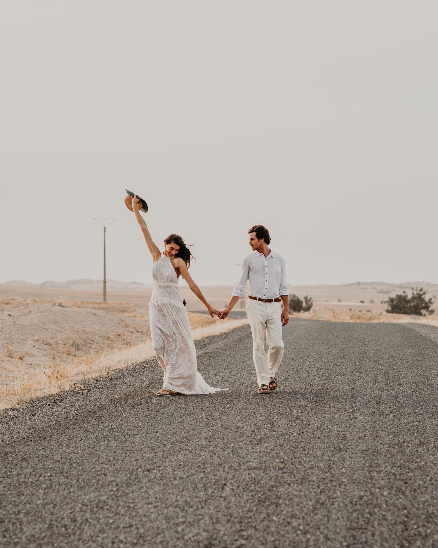A couple in white clothing holds hands while walking on a desert road with hat raised in celebration.