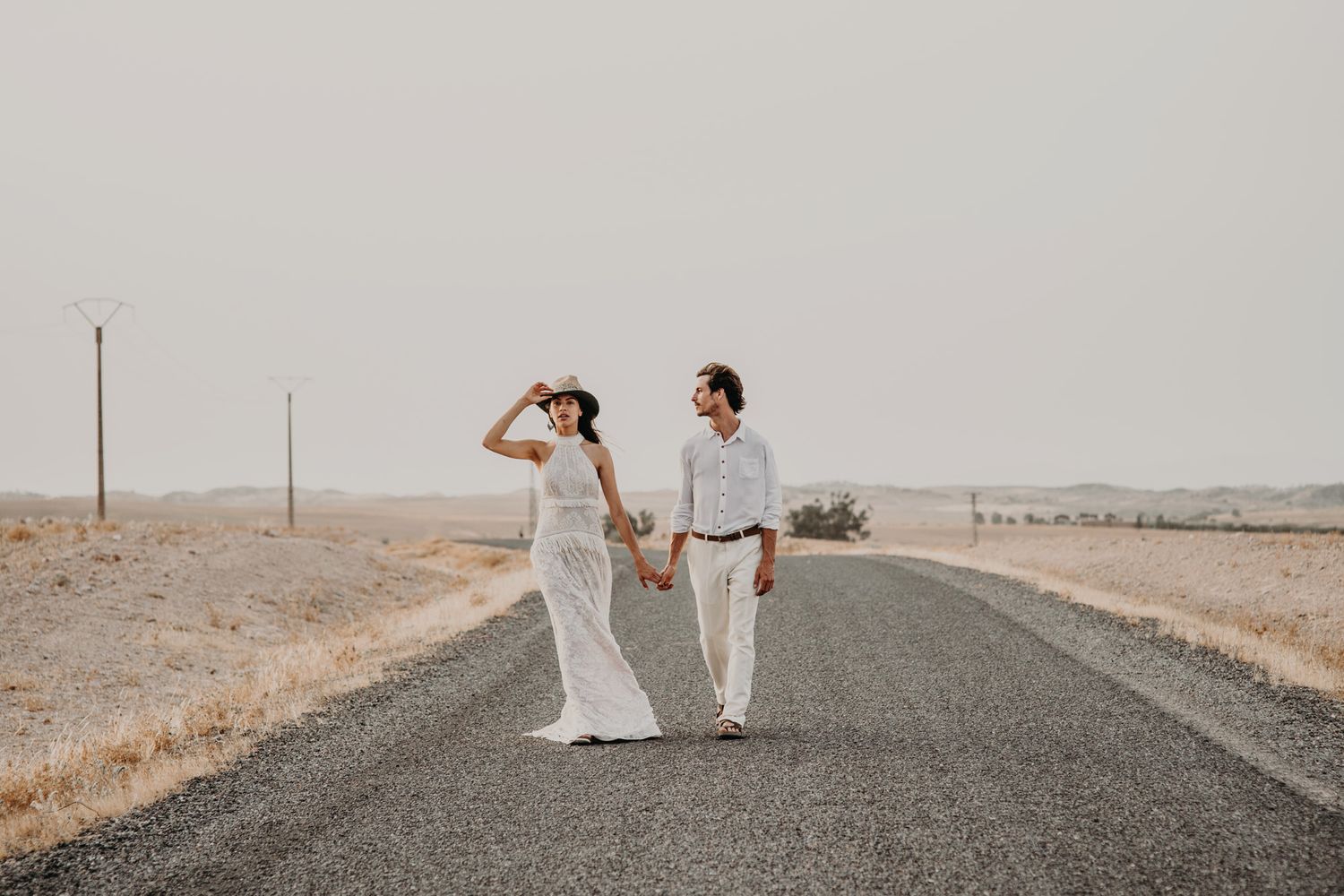A couple in white clothing holds hands while walking down a desert highway.