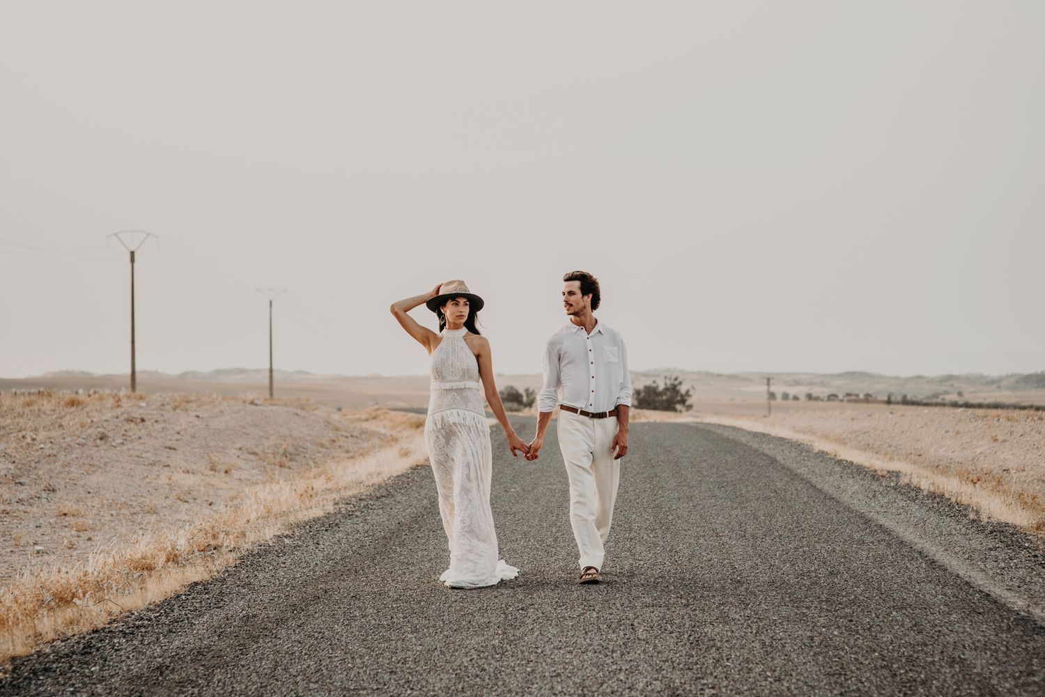 A couple in matching white outfits walks together on an empty desert road.