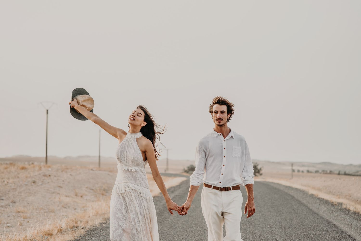 A playful desert wedding photo with a couple raising a hat while holding hands.