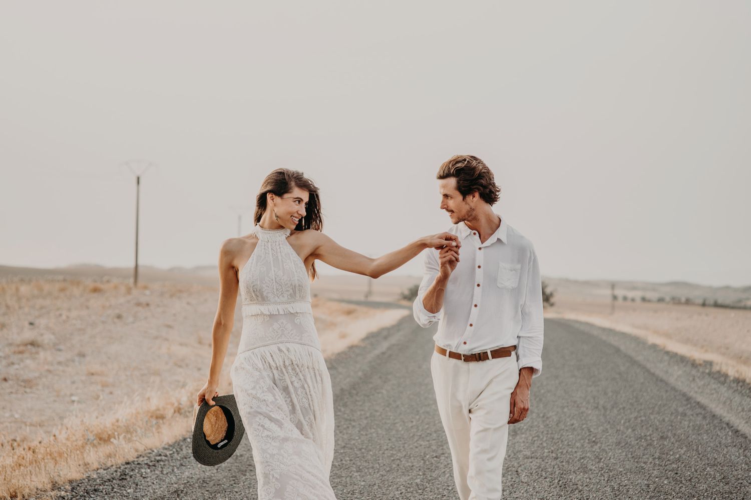 A couple in white outfits shares a playful moment with a hat on a desert road.
