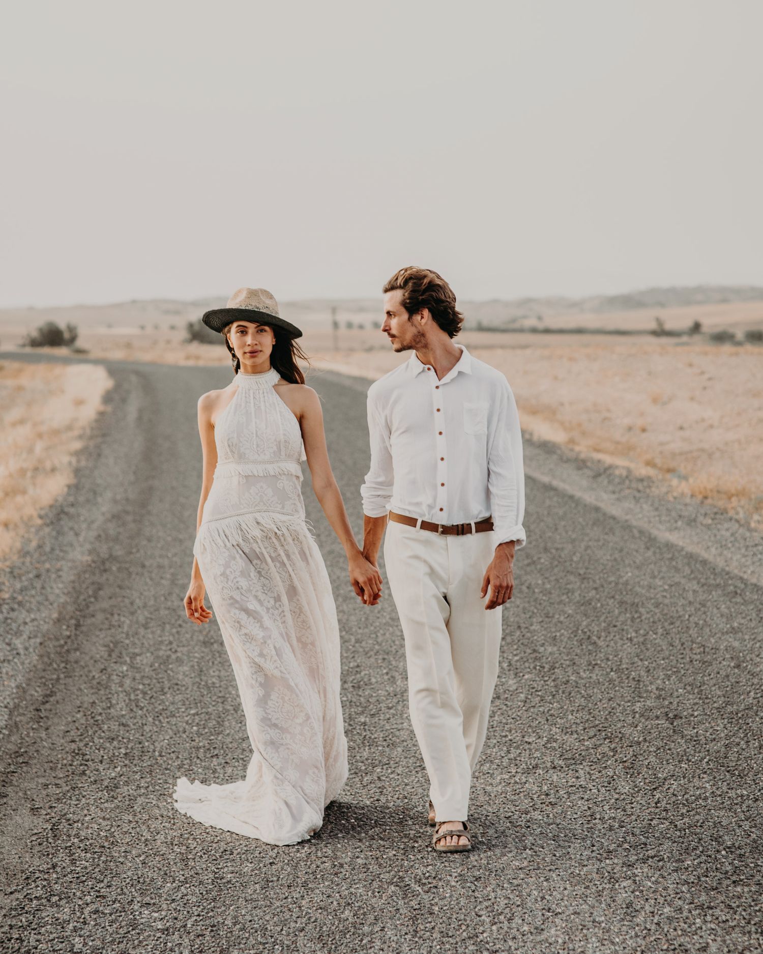 A stylish couple in white clothing walks hand in hand down a desert road.