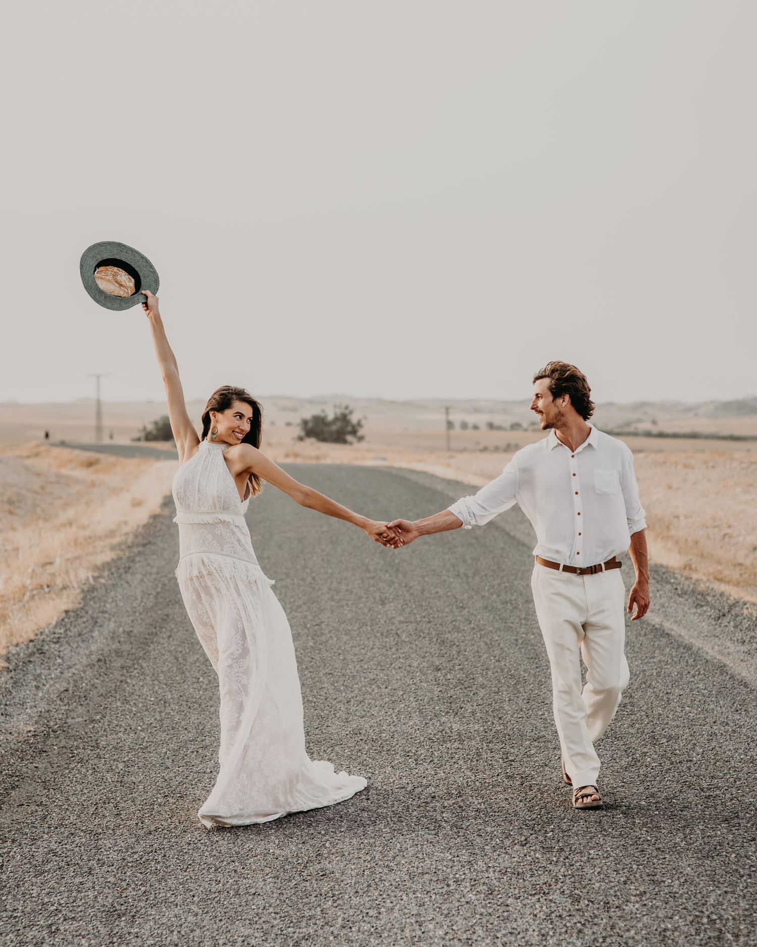 A couple in white attire plays with a hat while holding hands on a desert road.
