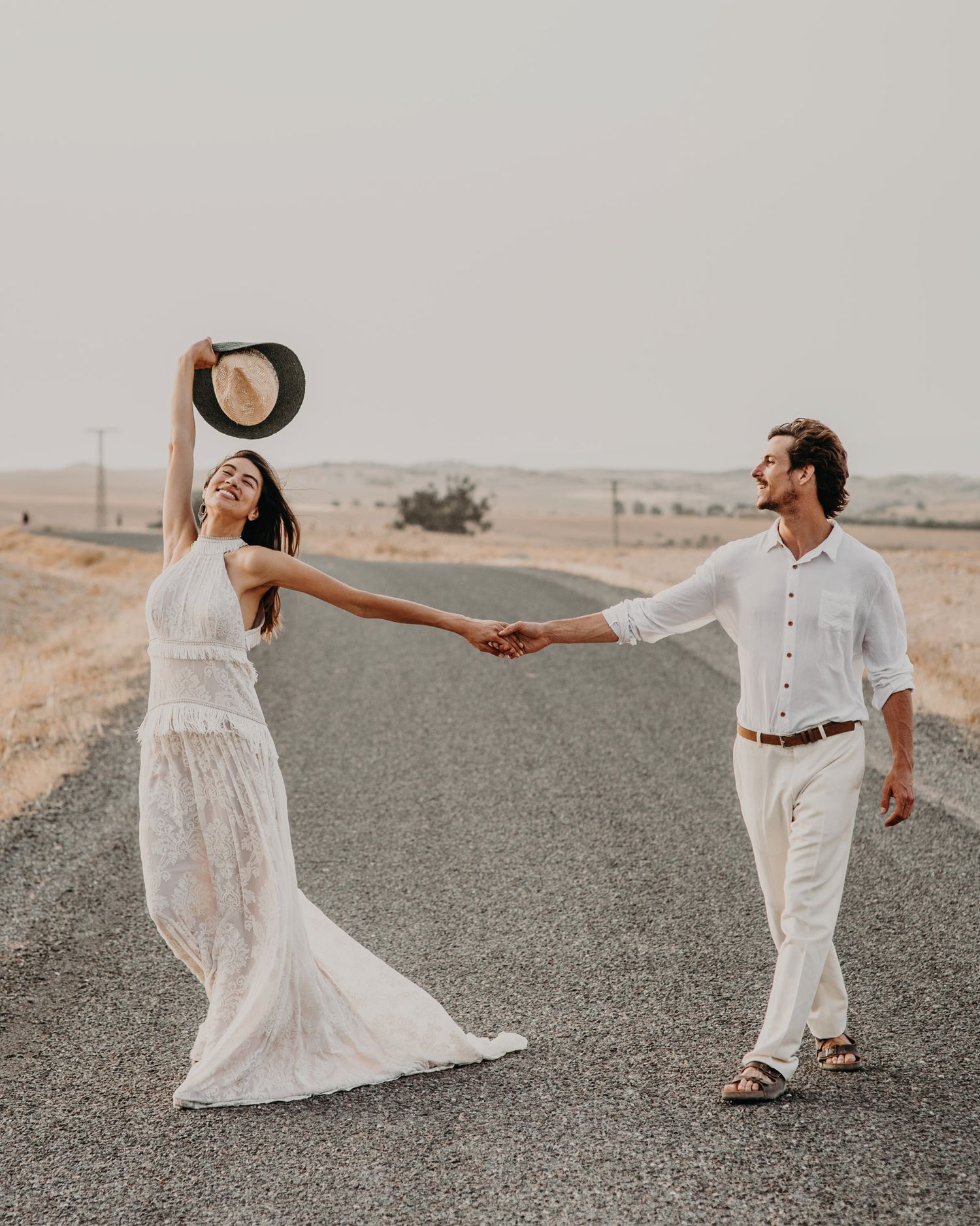 A couple in white clothing holds hands while dancing on an empty desert road.