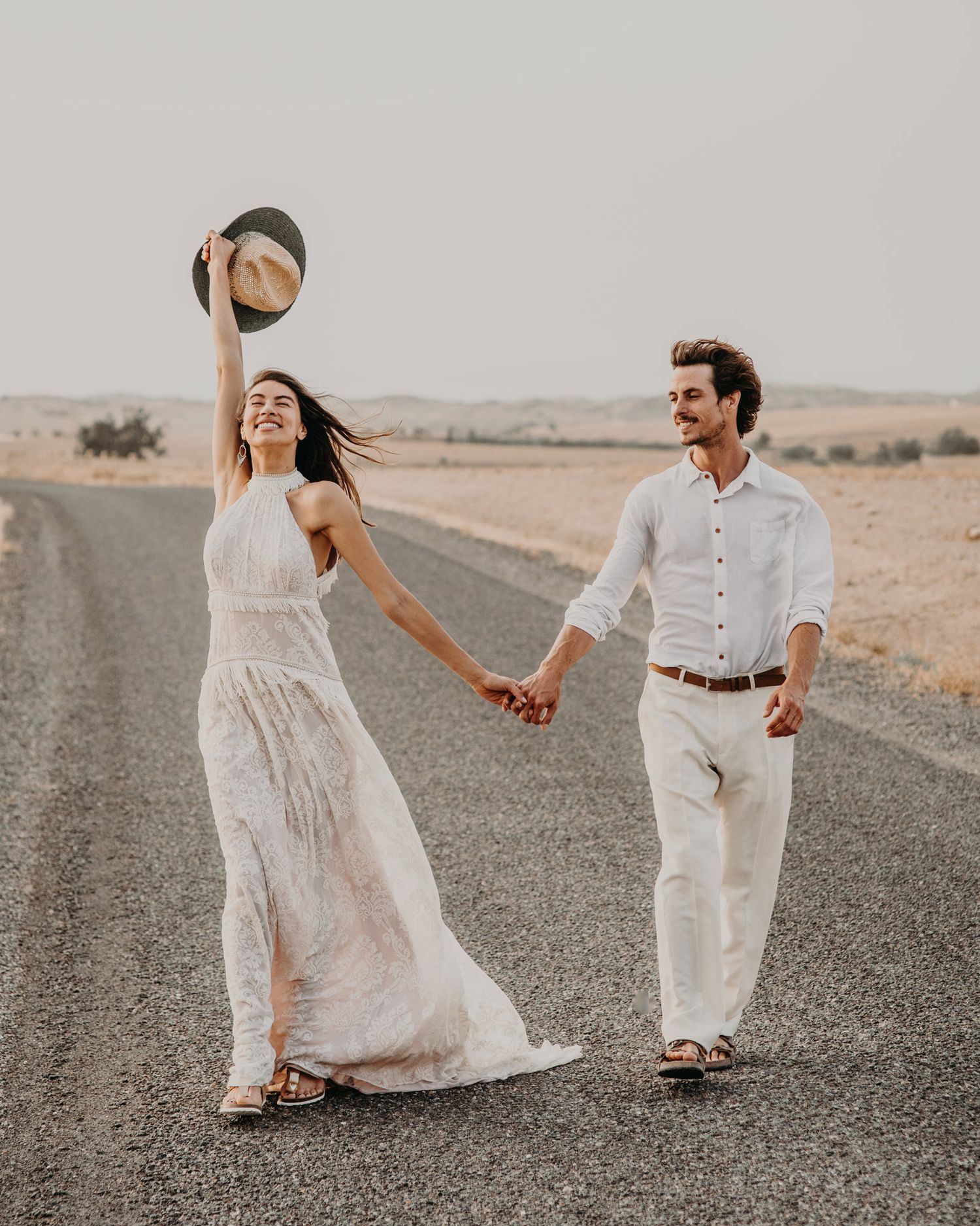 A couple in white outfits holds hands on a desert road while the woman playfully raises her hat.