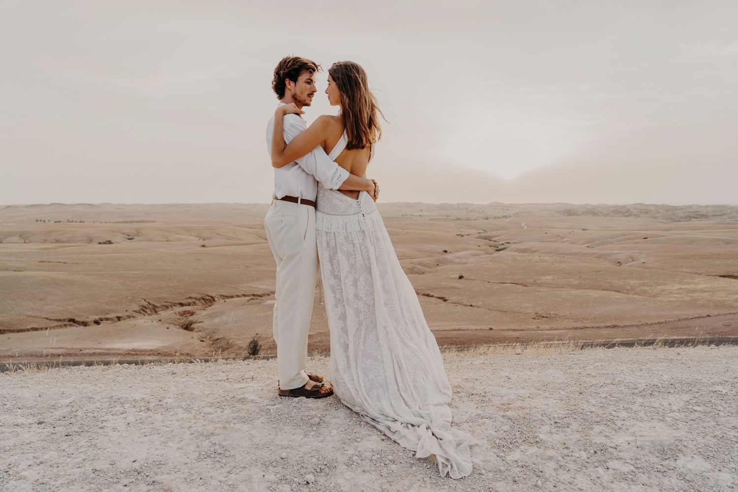 A romantic desert photoshoot featuring a couple in flowing white outfits sharing an intimate moment.