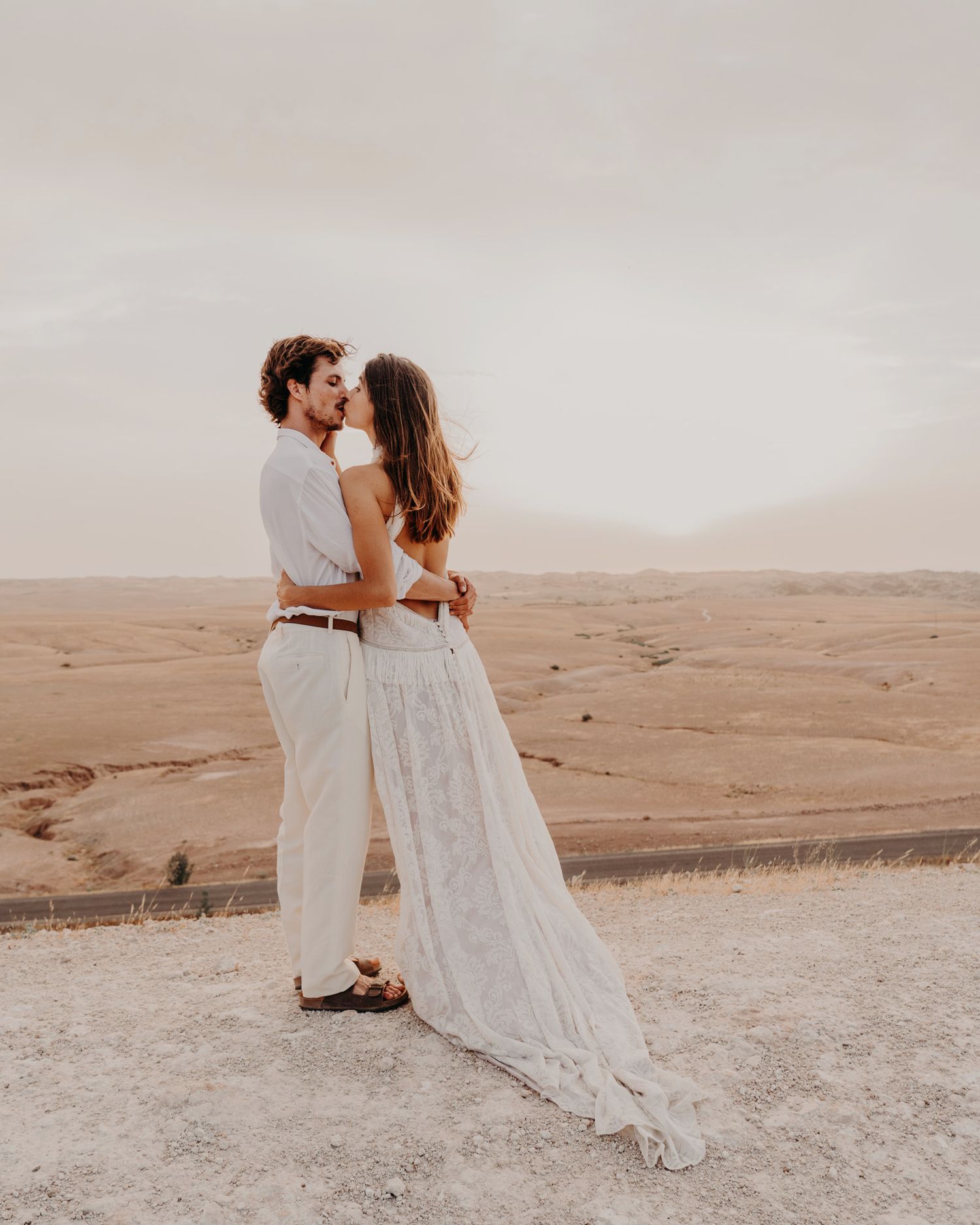 Couple embraces against a desert landscape backdrop wearing coordinated white attire.