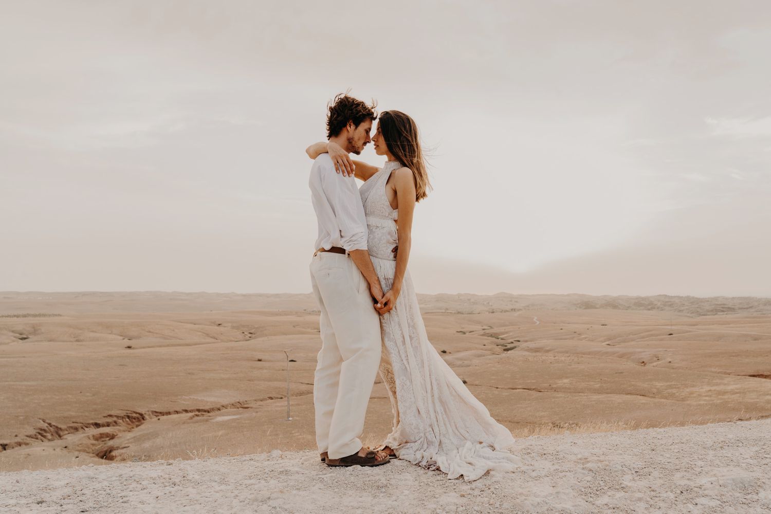 A dreamy desert wedding photo captures a couple in white clothing sharing a tender moment.