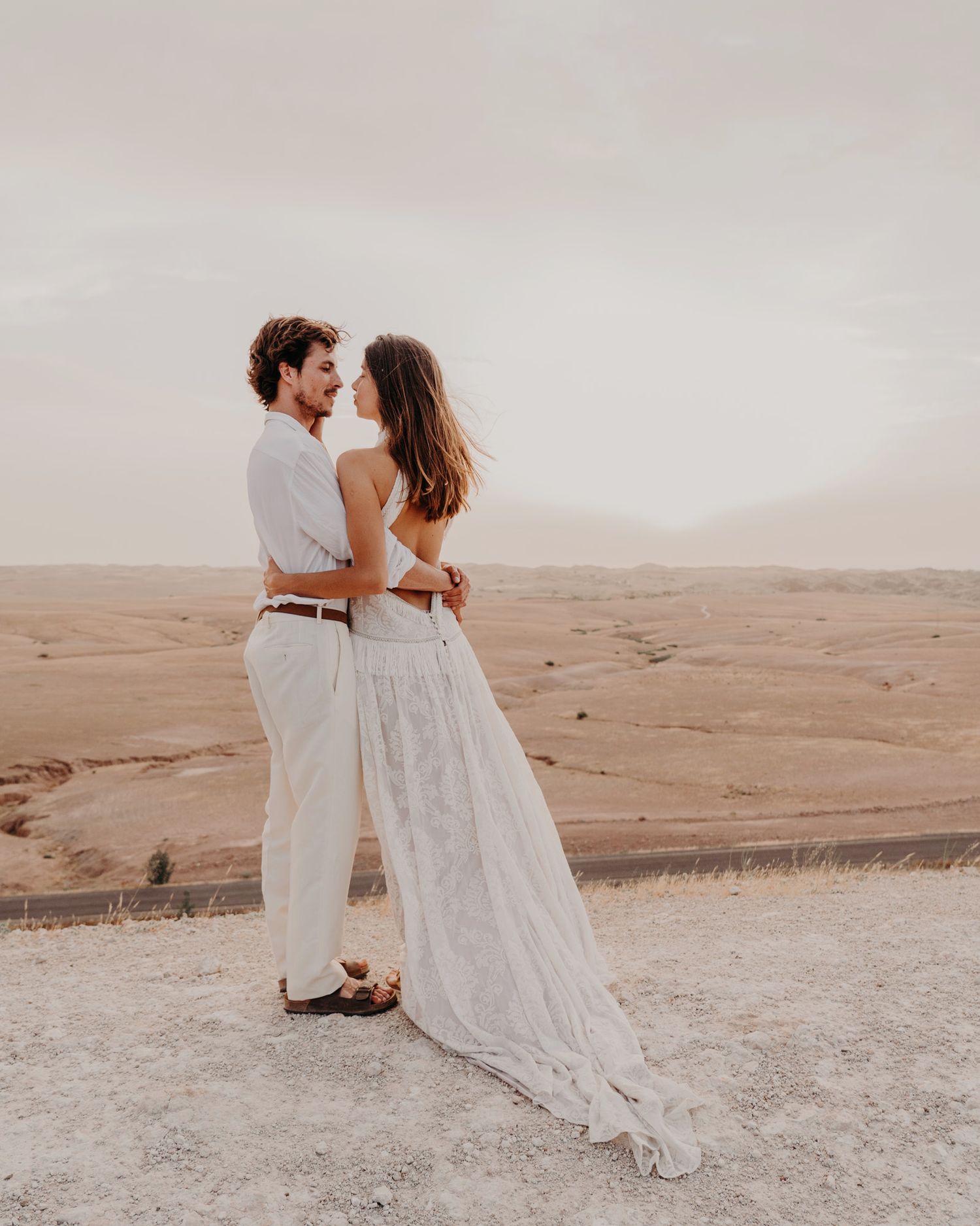 A romantic desert portrait shows a couple in white embracing against sandy hills.