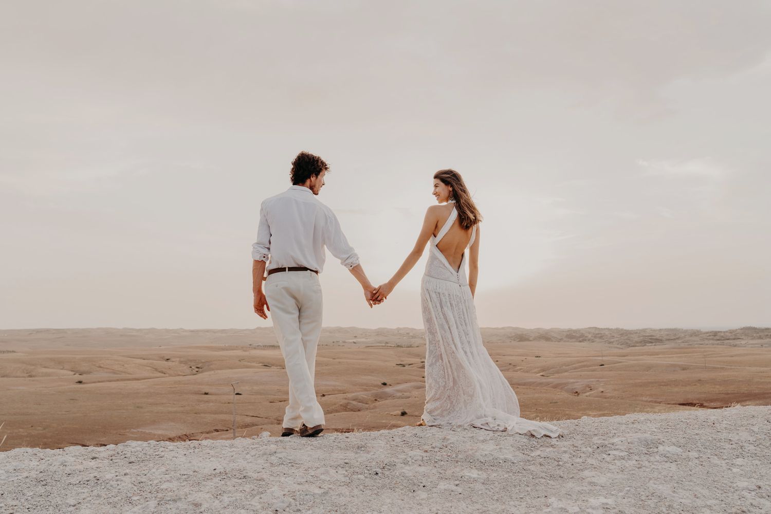 Couple in elegant white attire holds hands while walking in a desert setting.