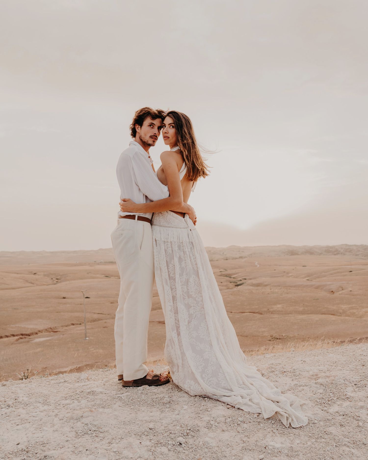 An intimate desert photo session features a couple in white clothing embracing.