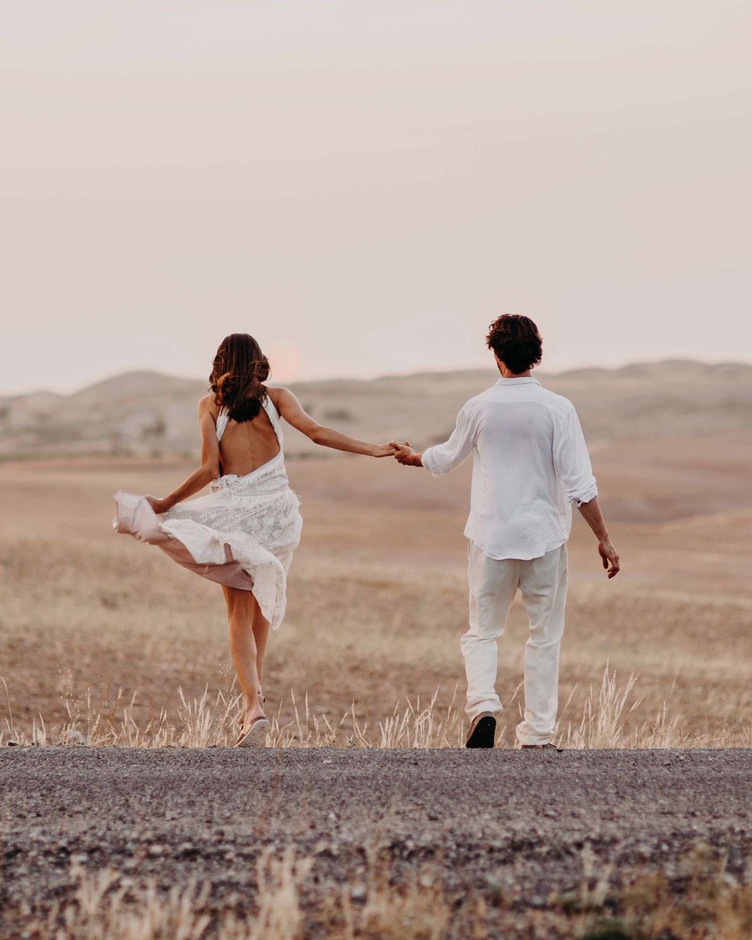 Two people in white clothing dance together on a dusty desert road at sunset.
